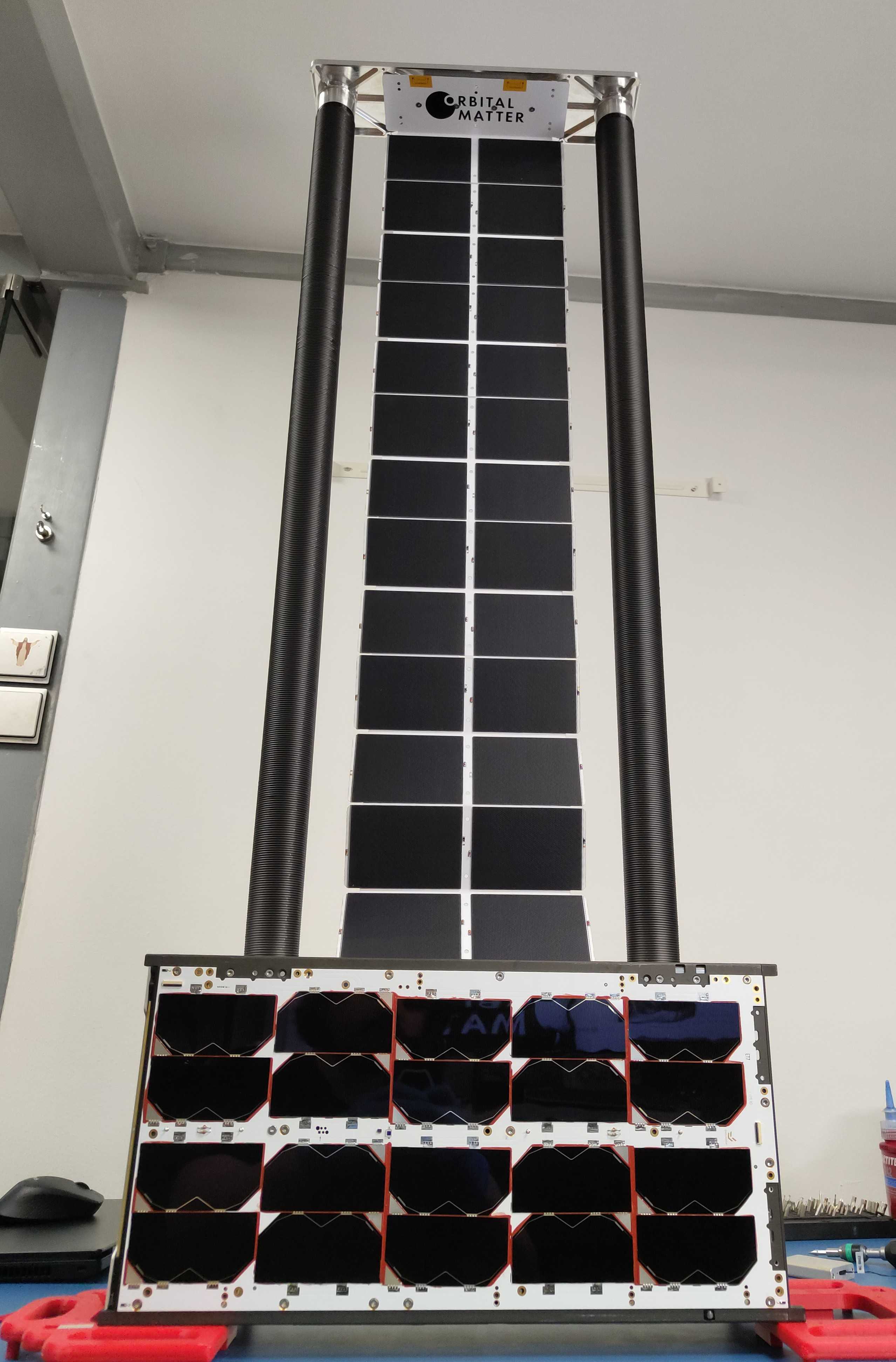 A flexible solar array deployed on a workbench by two 3D printed booms, with the folded array module mounted below.
