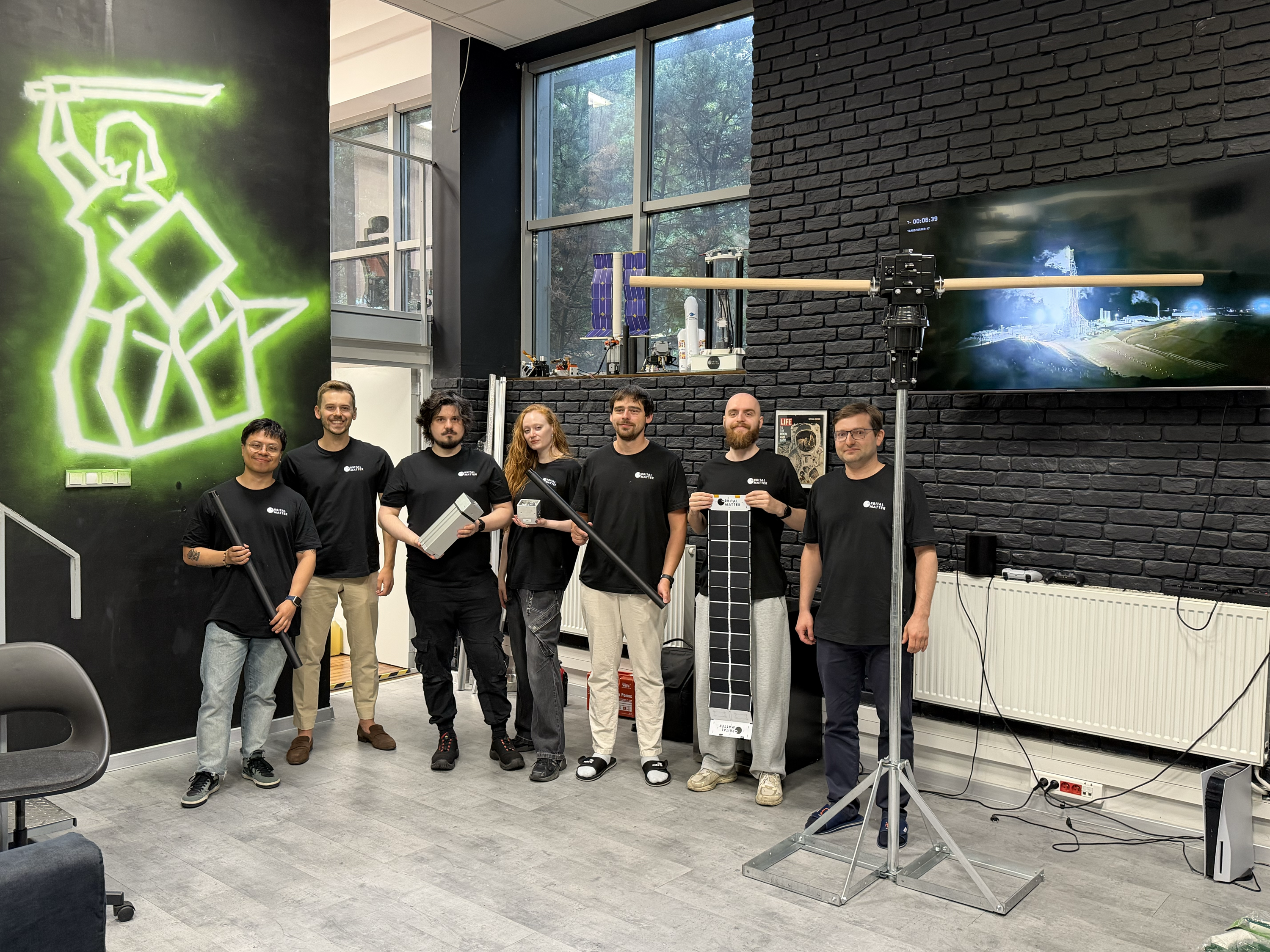 The Orbital Matter team standing together in their Warsaw office, holding printed booms, a CubeSat, and a solar array demonstrator.